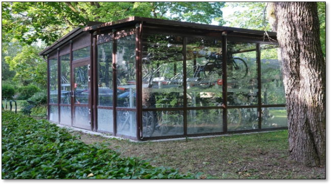 Foto som viser et større utendørs sykkelskur, der syklene er innelåst med adgangskontroll. Dette sykkelskuret er gjennomsiktig og har parkering innvendig i to etasjer, der det står flere sykler parkert.