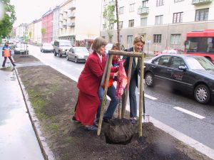 Fotografi av tre personer som planter et løvtre ved siden av en trafikkert bygate.