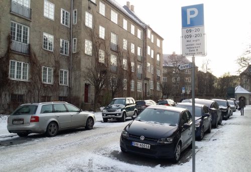 Biler parkert langs en bygate – 4 etasjes bygård i bakgrunnen.