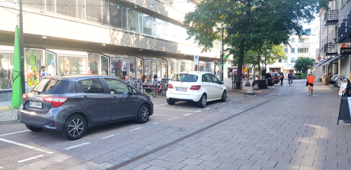 Biler parkert i markerte områder langs en vei i et by/sentrumsmiljø.