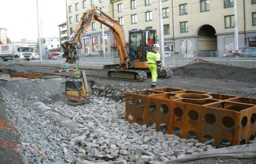 Fotoet viser en kranbil som løfter på plass støpejernskummer ved siden av veien.